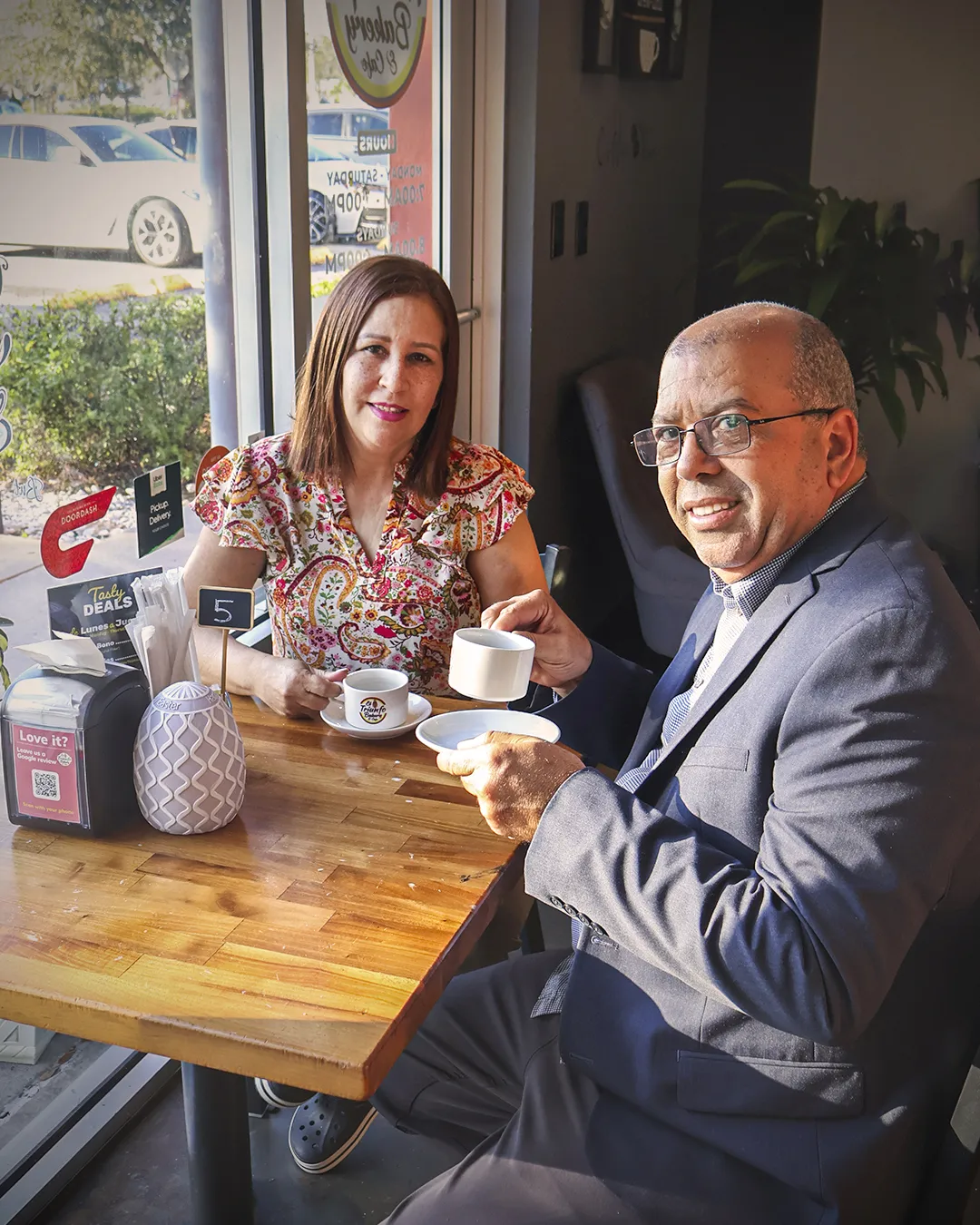 92a49787-e767-426a-b4b4-beaa50fbaa3e.webp Smiling man and woman enjoying coffee at a sunny cafe table
