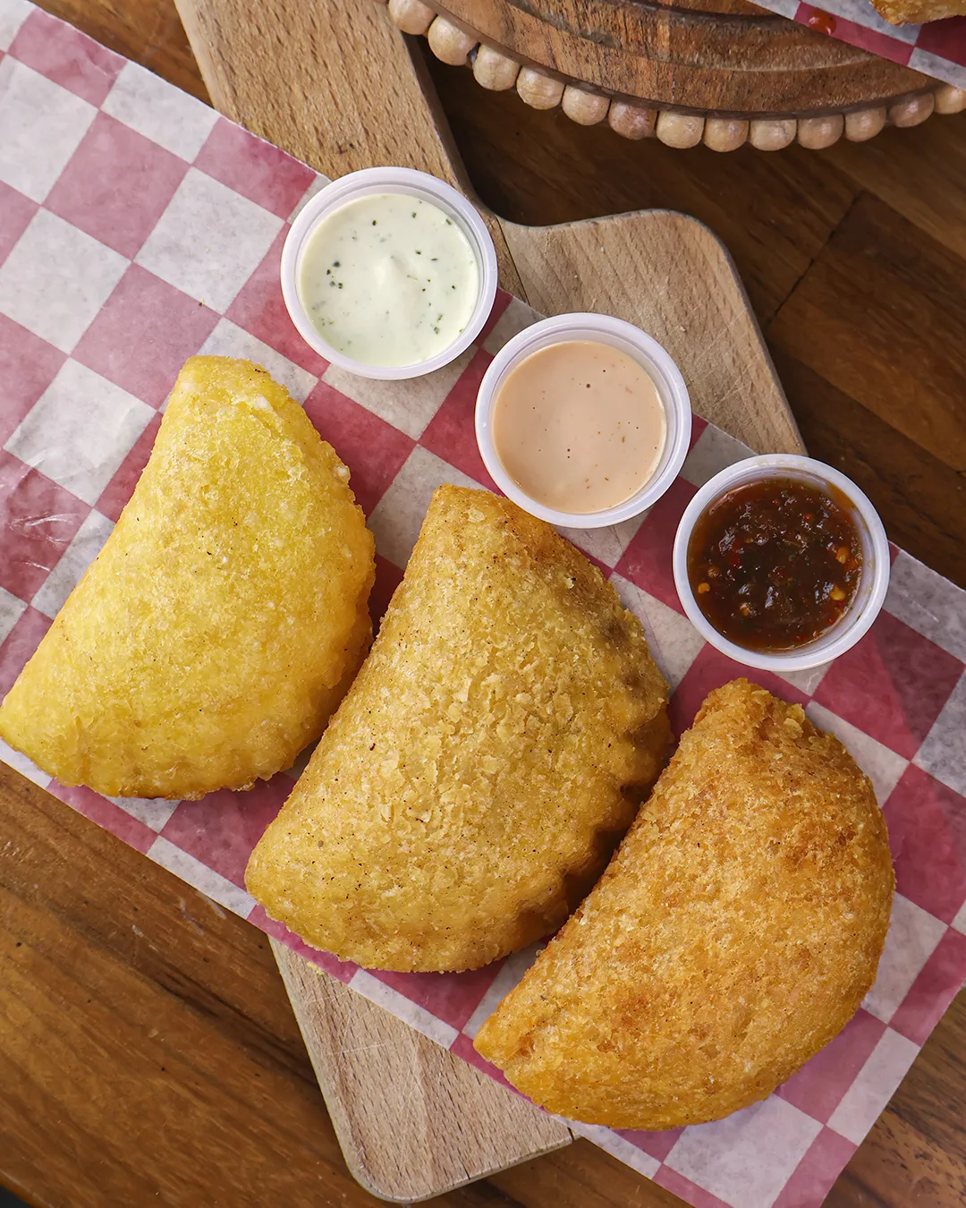 e89ea8cf-6b73-48fc-be1b-54ad13606f46.webp Three fried empanadas on checkered paper with white, beige, and red sauces
